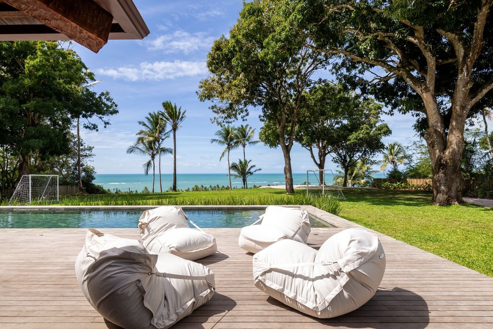PISCINA | Na área externa, a piscina se estende diante do gramado e da vista para o mar. Sobre o deque de madeira, os pufes da HIO Decor criam um espaço descontraído para relaxar e aproveitar a paisagem — Foto: Antonio Soto/Divulgação