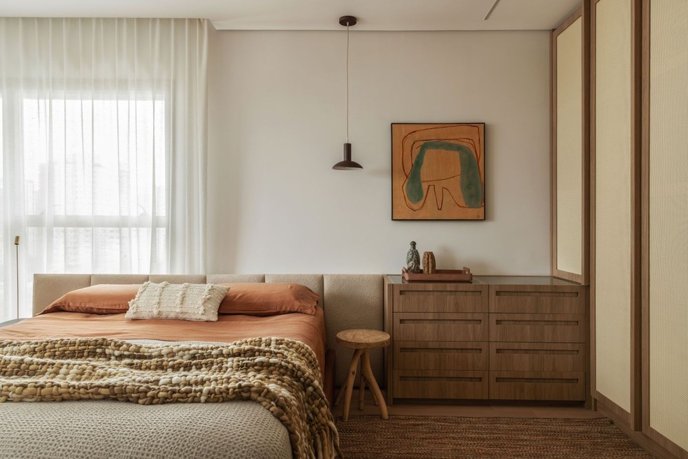 A cama em marcenaria revestida de couro e cabeceira estofada, com execução da Bossa Estofados, recebeu enxoval da Casa Almeida e almofada branca da Codex Home. Sobre o banquinho da loja Mau, que serve de apoio lateral, está a luminária pendente Dri, da Golden Art. Ao lado, móvel feito de MDF Itapuã, da Duratex, mesmo material do guarda-roupa, que recebeu portas de palha indiana natural quadriculada. Quadro da Dpot Objeto. Tapete da Botteh — Foto: Maura Mello/Divulgação | Projeto do escritório TODOS Arquitetura
