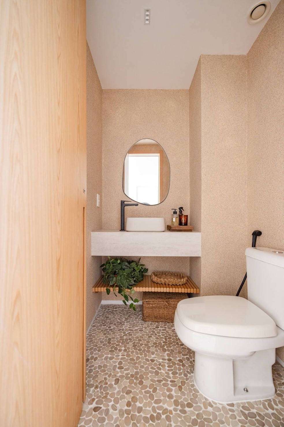 LAVABO | Com atmosfera de spa, o lavabo combina piso de seixos e paredes com textura de efeito areia, da I Colori di Venezia. A cuba de apoio é de concreto e a bancada, de lâmina ultracompactada. A marcenaria em lâmina de carvalho-americano acompanha os metais e a louça da Deca — Foto: Pedro Mello/Interior da Foto/Divulgação
