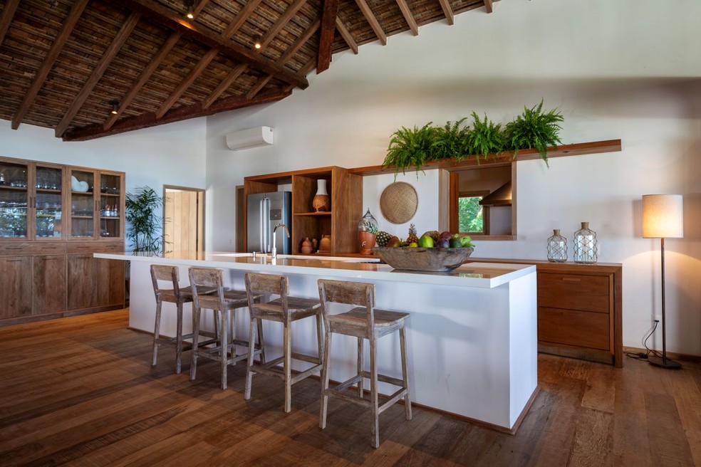 COZINHA | Ao lado da sala de jantar, a cozinha tem a ilha branca que contrasta com a marcenaria fixa de madeira, desenhada pelo escritório e executada em parceria com a Arte Baiana. Banquetas rústicas acompanham a bancada, enquanto o piso de madeira de demolição, fornecido por Lucas Vítor, de Tiradentes, MG, remete à linguagem dos interiores — Foto: Antonio Soto/Divulgação